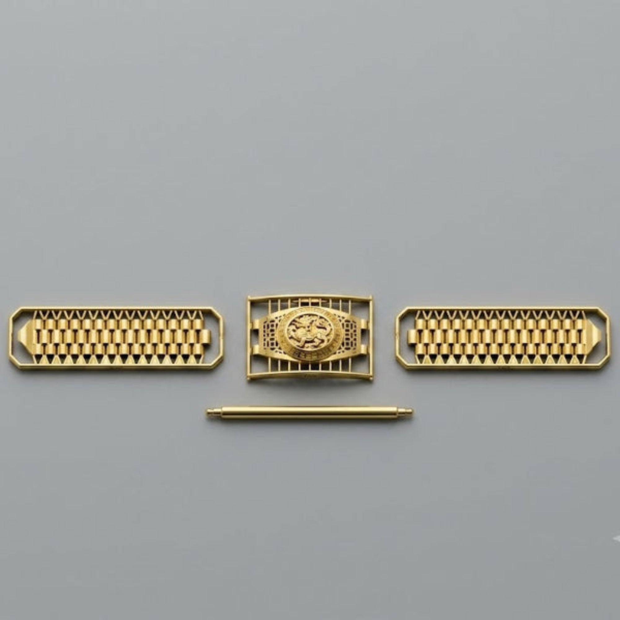 A set of gold-toned mechanical watch components or decorative hardware pieces arranged horizontally on a plain gray background, including two rectangular grids with small circular elements and a central rectangular piece with a circular emblem, plus a thin horizontal rod below.