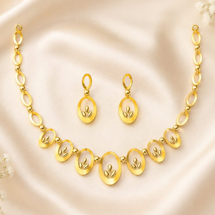 Gold necklace and matching drop earrings laid on a soft cream satin background with small white flowers at the corners. The jewelry features oval links and oval pendant motifs with a raised leaf design in the center.