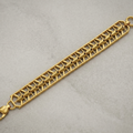 A gold-toned metal link bracelet with a geometric, openwork trellis pattern, featuring a lobster clasp and a top loop, shown on a textured light gray background.