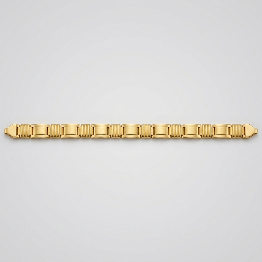 A gold tennis bracelet with a distinctive fluted or ribbed link design, displayed horizontally against a clean, light gray gradient background.