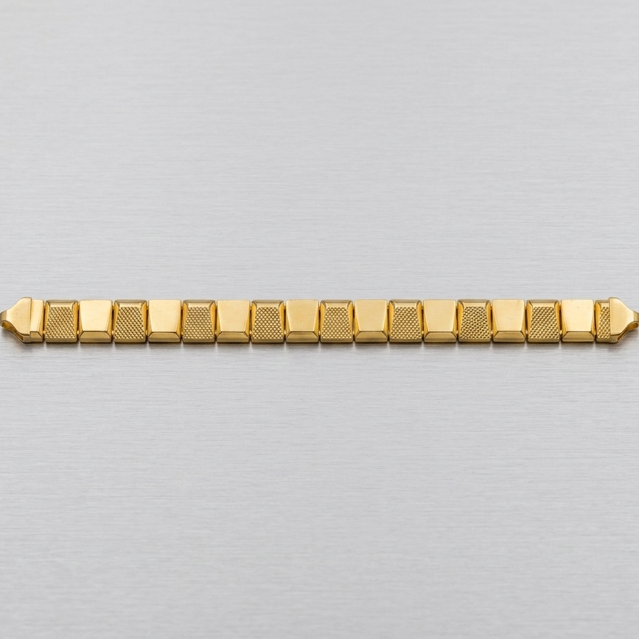 A gold-toned metal link bracelet with a textured pattern on the links, displayed horizontally on a brushed silver metal surface.