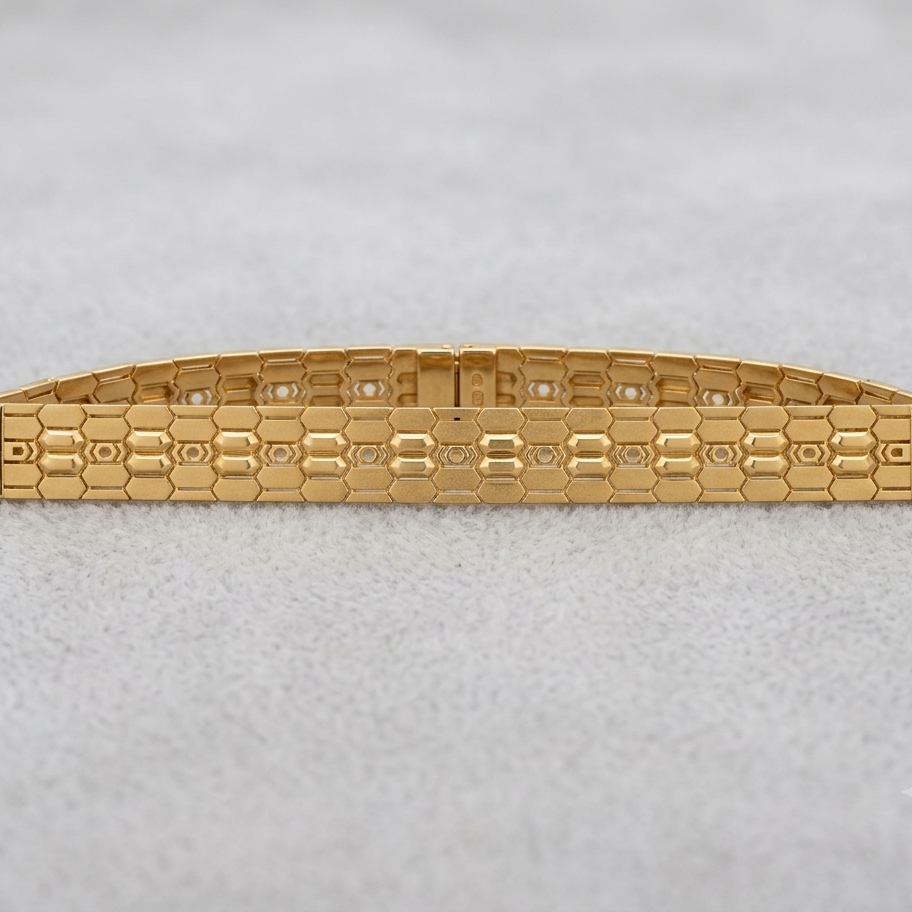 A gold-tone metal bracelet with a textured honeycomb pattern and small clear stones set along the top, displayed on a light gray textured background.