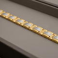 A close-up of a gold-tone metal link bracelet with textured silver inlay panels, resting diagonally on a dark gray textured surface.
