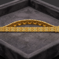 A gold bracelet with a textured, geometric link design, displayed on a dark gray velvet jewelry box.