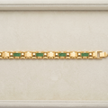 A gold link bracelet with green and pink pavé accents, displayed horizontally on a light beige textured jewelry box.