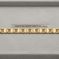 A gold-colored metal watch band with a repeating rectangular link pattern, displayed horizontally on a gray textured surface with a small label above it.