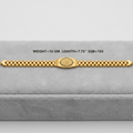 A gold bracelet displayed on a gray velvet jewelry box. The bracelet features a central textured oval clasp and a beaded link design.