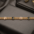 A gold-toned bracelet with alternating square pinkish-beige and black enamel panels, each featuring an Om symbol, displayed on a dark gray satin surface.