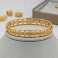 A gold bracelet displayed on a light gray jewelry bust, with matching gold earrings and a jewelry box blurred in the background.