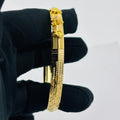 A gold-plated bangle bracelet with detailed engravings and a decorative clasp, held by a hand wearing a black glove against a plain light gray background.