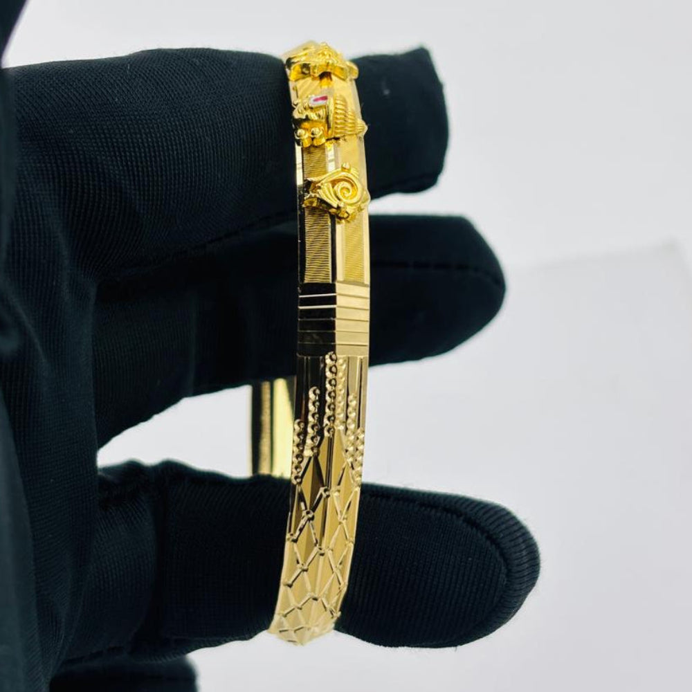 A gold-plated bangle bracelet with detailed engravings and a decorative clasp, held by a hand wearing a black glove against a plain light gray background.