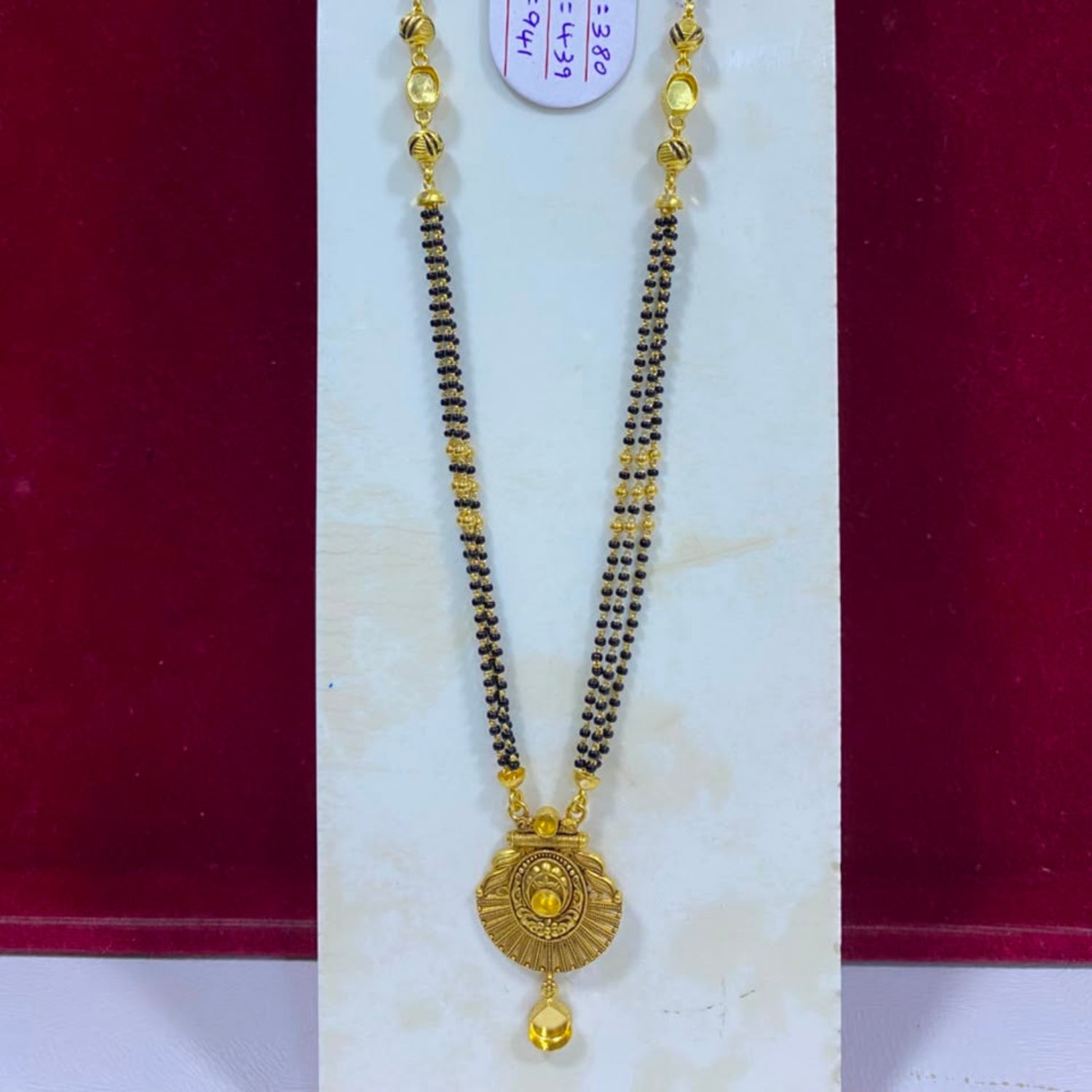 A gold and black beaded necklace with a round gold pendant, displayed on a white jewelry stand against a dark red background.