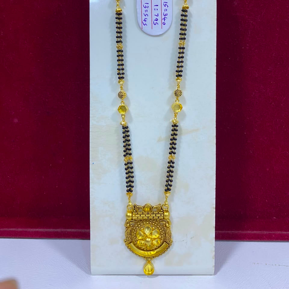 A traditional Indian gold necklace with a large ornate gold pendant and a long black bead chain with small gold accents, displayed on a white stand against a dark red background.