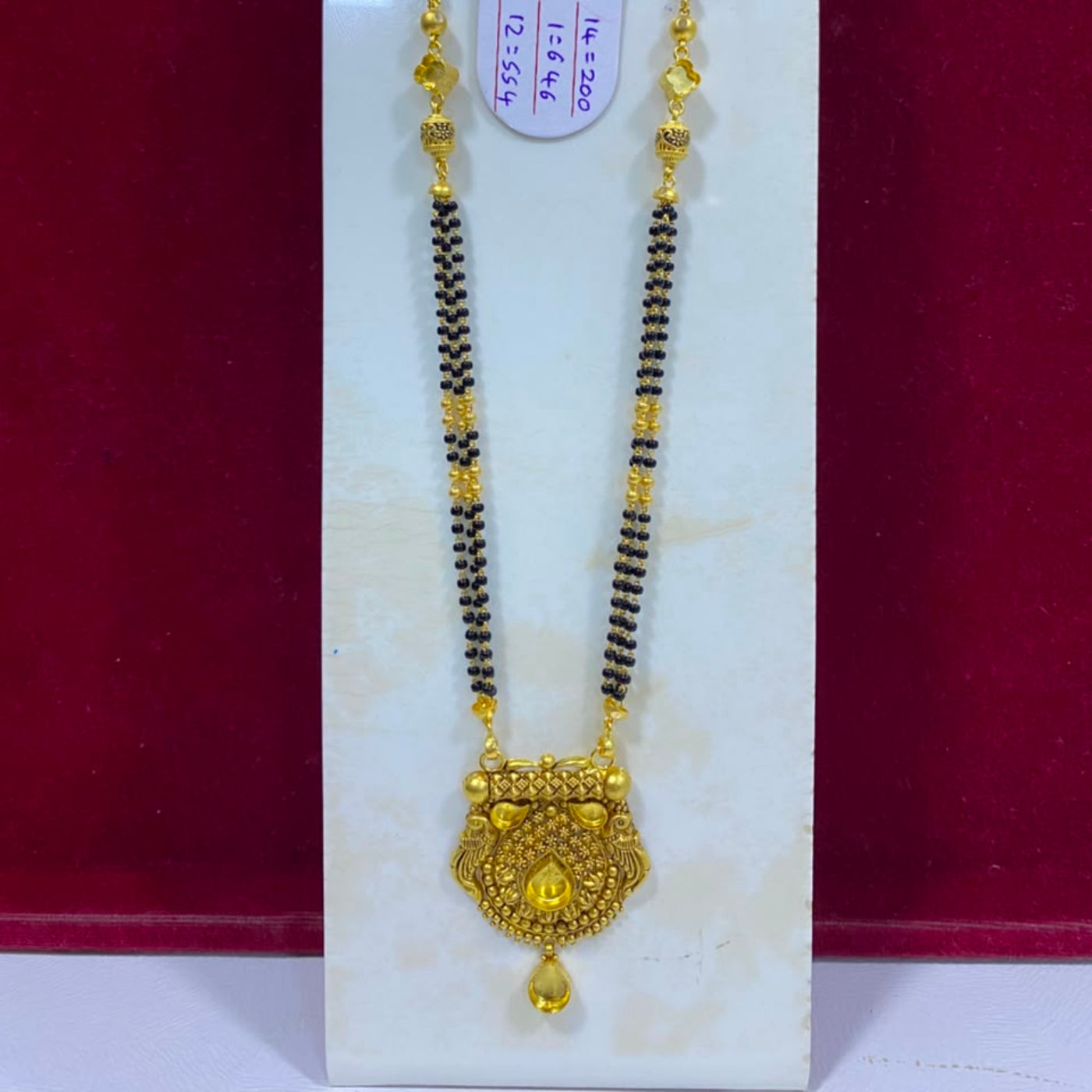 A gold and black beaded necklace with an ornate gold pendant, displayed on a white stand against a dark red background.