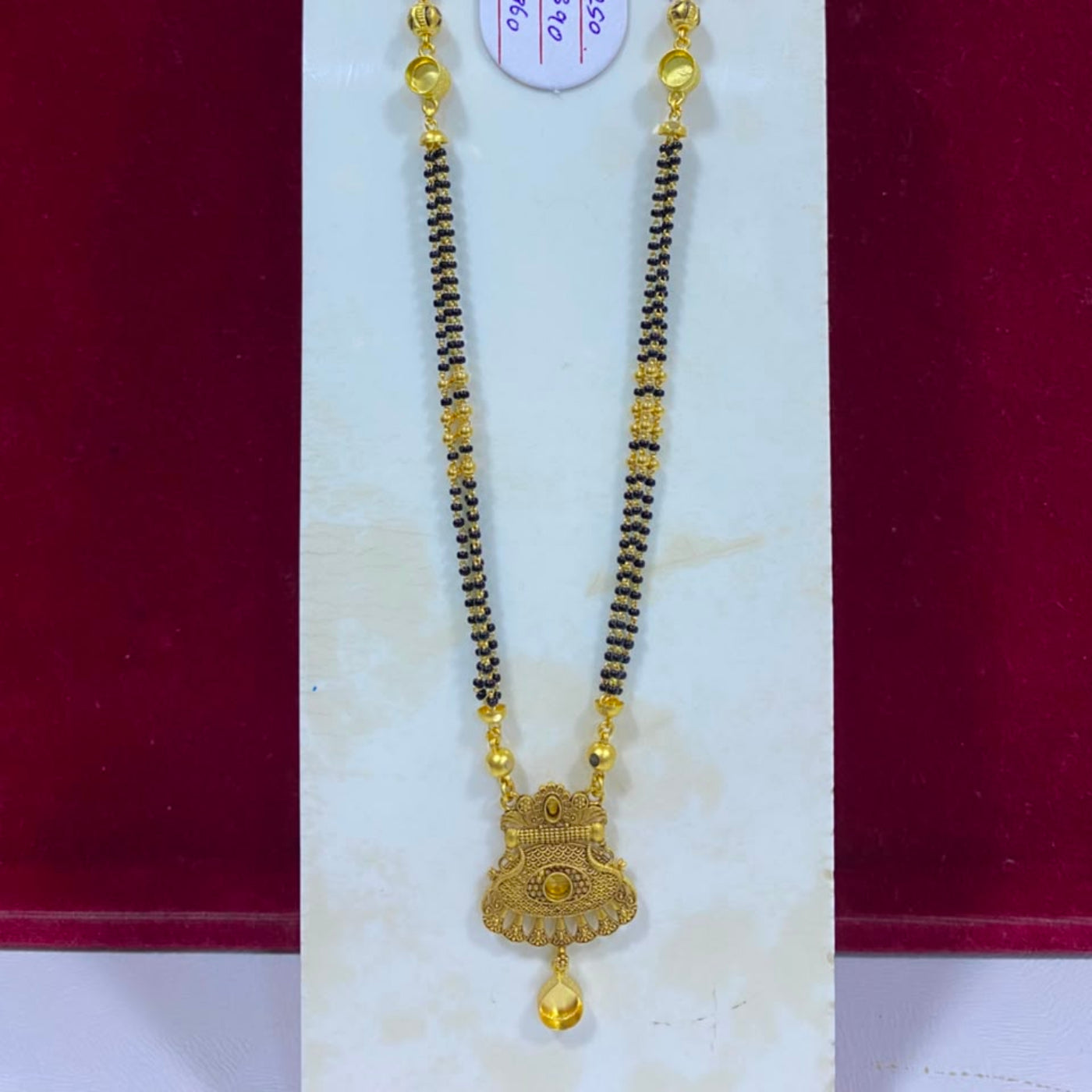 A gold and black beaded necklace with a decorative gold pendant, displayed on a white jewelry stand against a dark red background. A small white price tag is attached near the top.