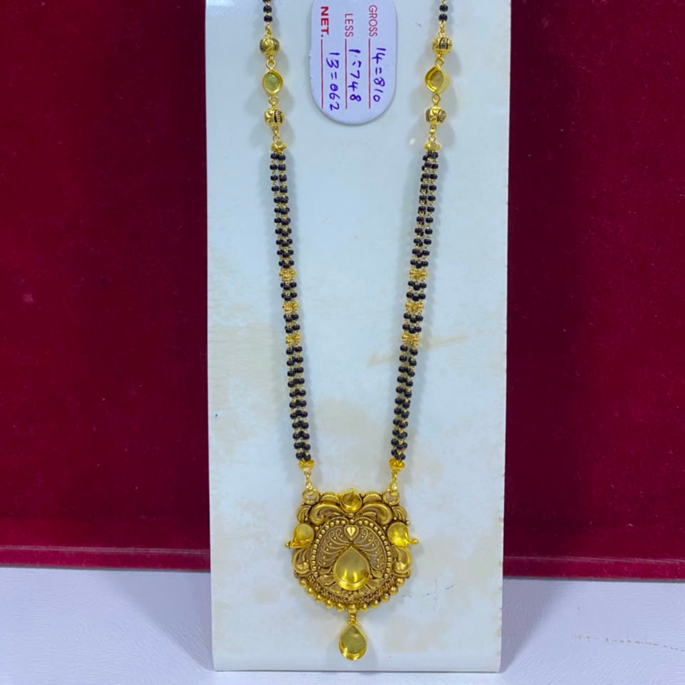 A gold and black beaded necklace with an ornate gold pendant, displayed on a white stand against a dark red background. A small white tag with handwritten measurements is attached to the necklace.