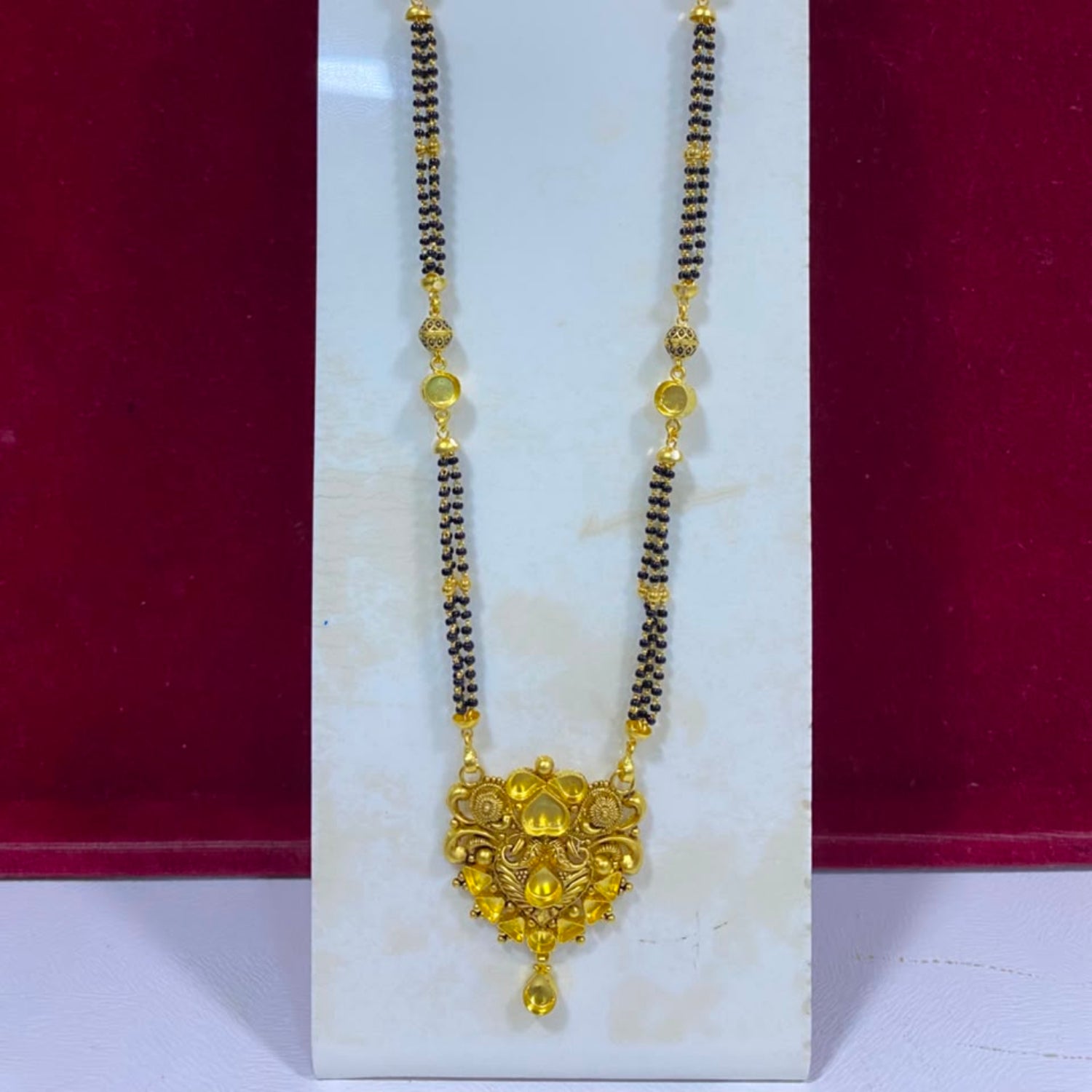 A traditional Indian gold and black bead necklace displayed on a white jewelry stand against a dark red background.