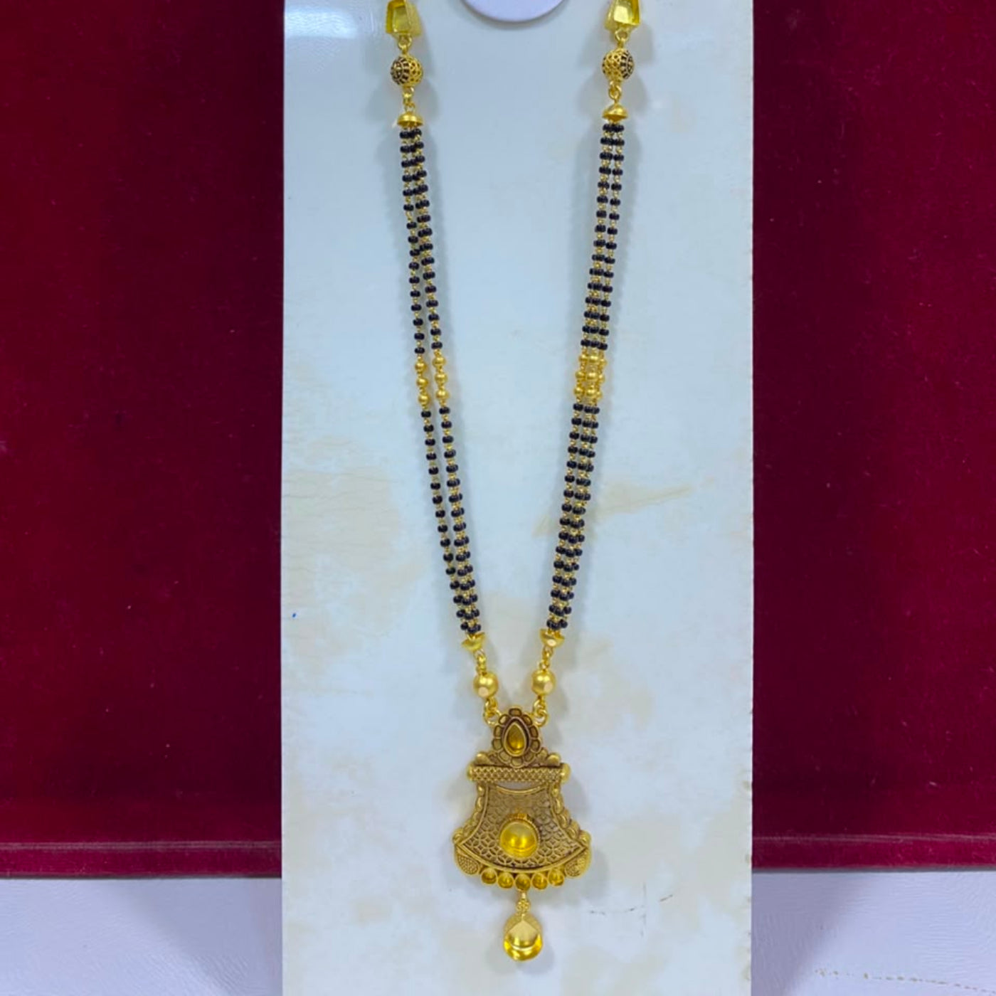 A traditional Indian gold and black beaded necklace (malari) with a decorative gold pendant, displayed on a white stand against a dark red background.
