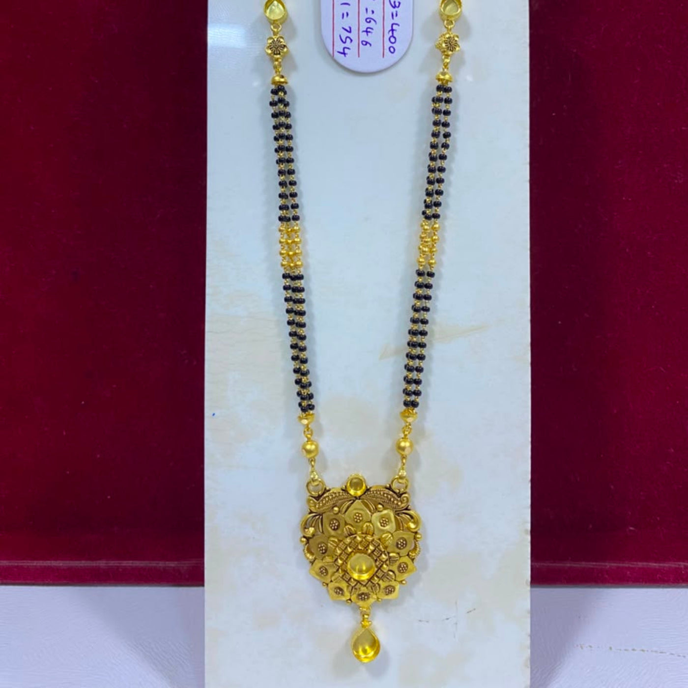 A gold and black beaded necklace with an ornate gold pendant, displayed on a white stand against a dark red background. A white product tag is attached to the necklace.