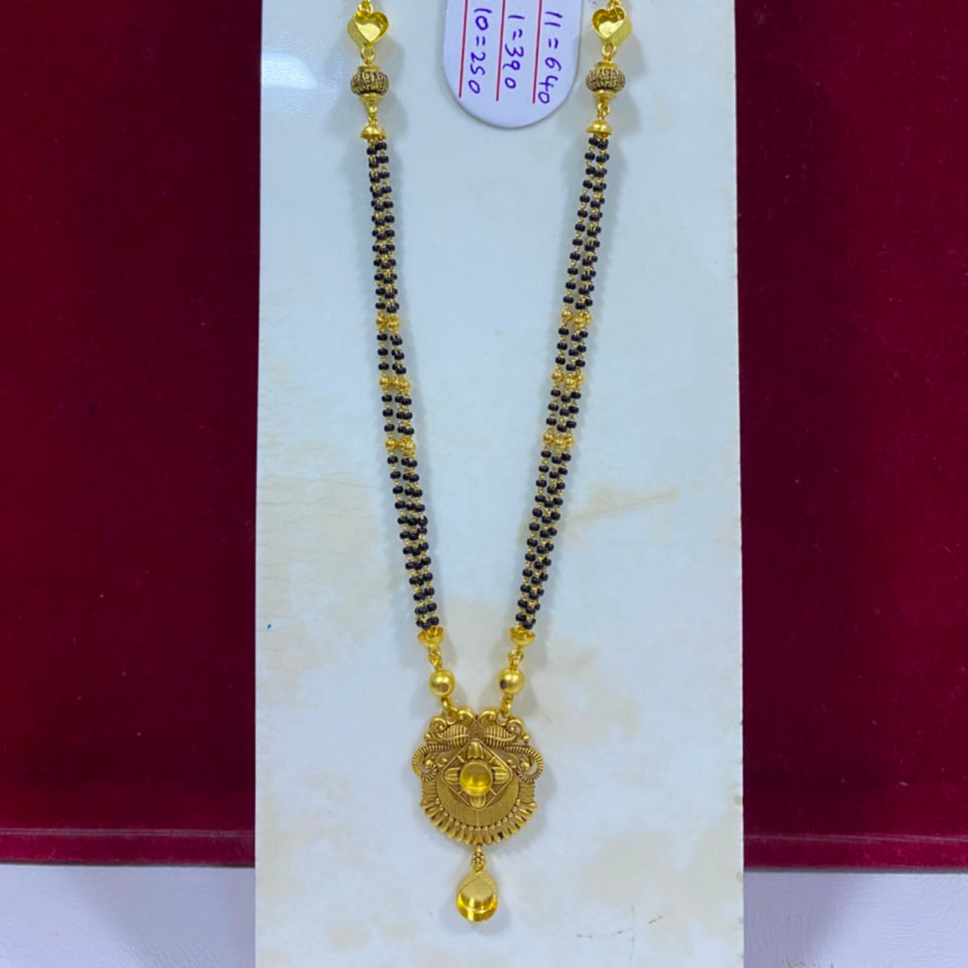 A gold and black beaded necklace with a detailed gold pendant, displayed on a white stand against a dark red background. A white price tag is attached to the necklace.