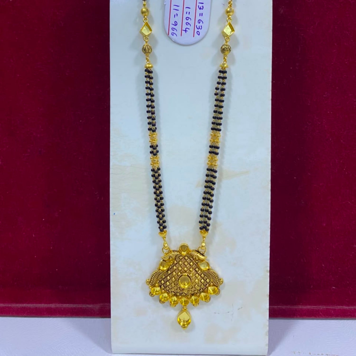 A traditional Indian gold and black bead necklace with a detailed gold pendant, displayed on a white stand against a dark red background.
