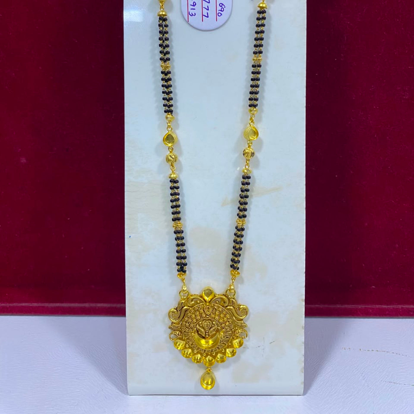 A gold and black beaded necklace with a detailed gold pendant, displayed on a white stand against a dark red background.