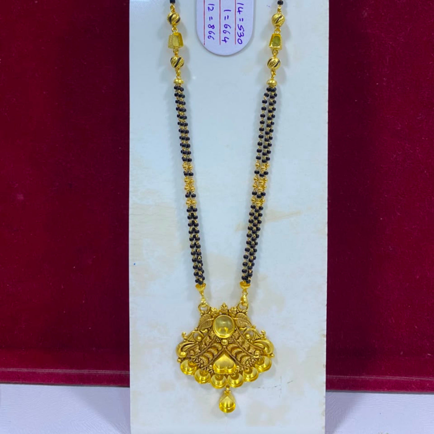 A gold and black beaded necklace with an ornate gold pendant, displayed on a white vertical stand against a dark red background. A small white tag with red text is attached near the top.