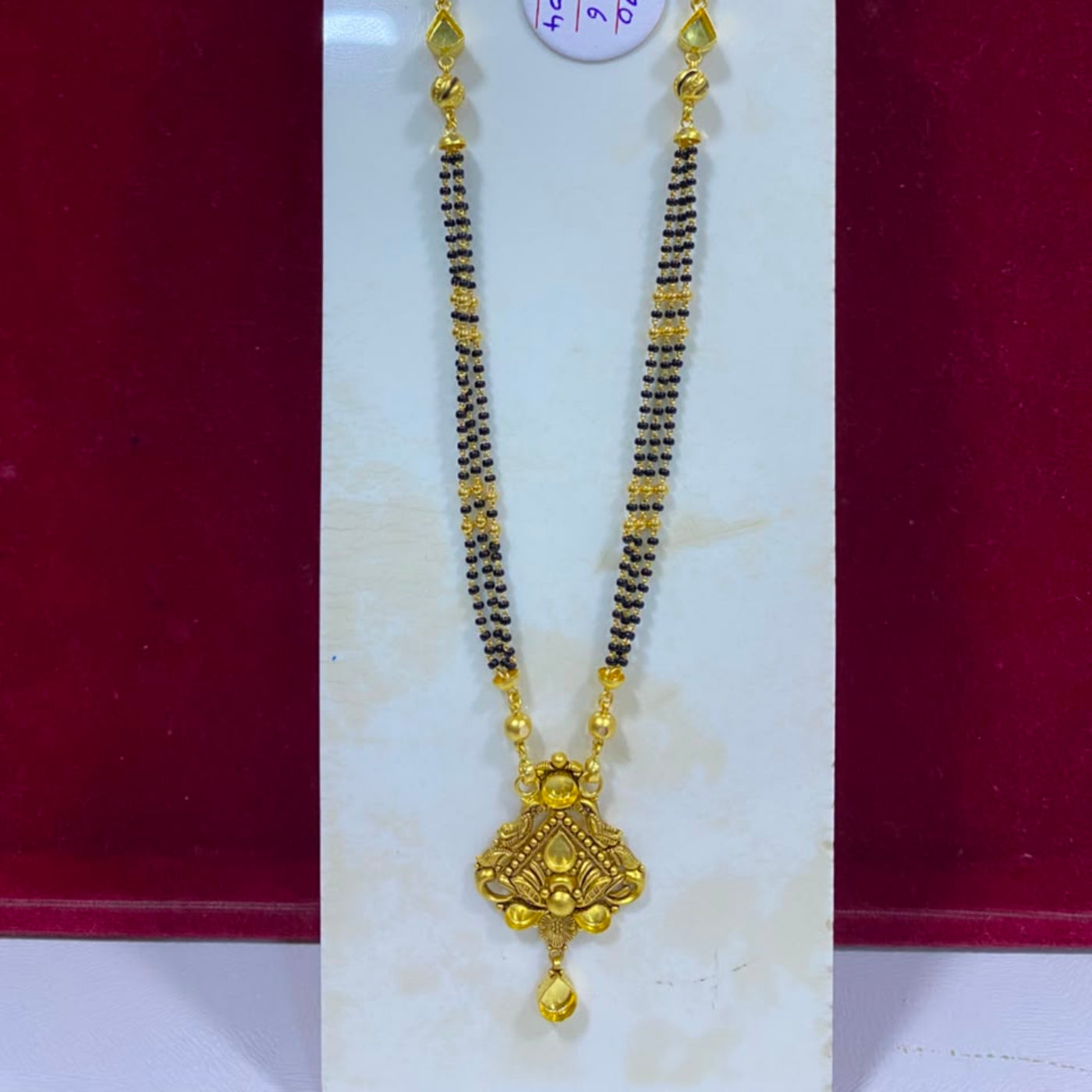 A traditional Indian gold and black beaded necklace with a detailed gold pendant, displayed on a white stand against a red background.