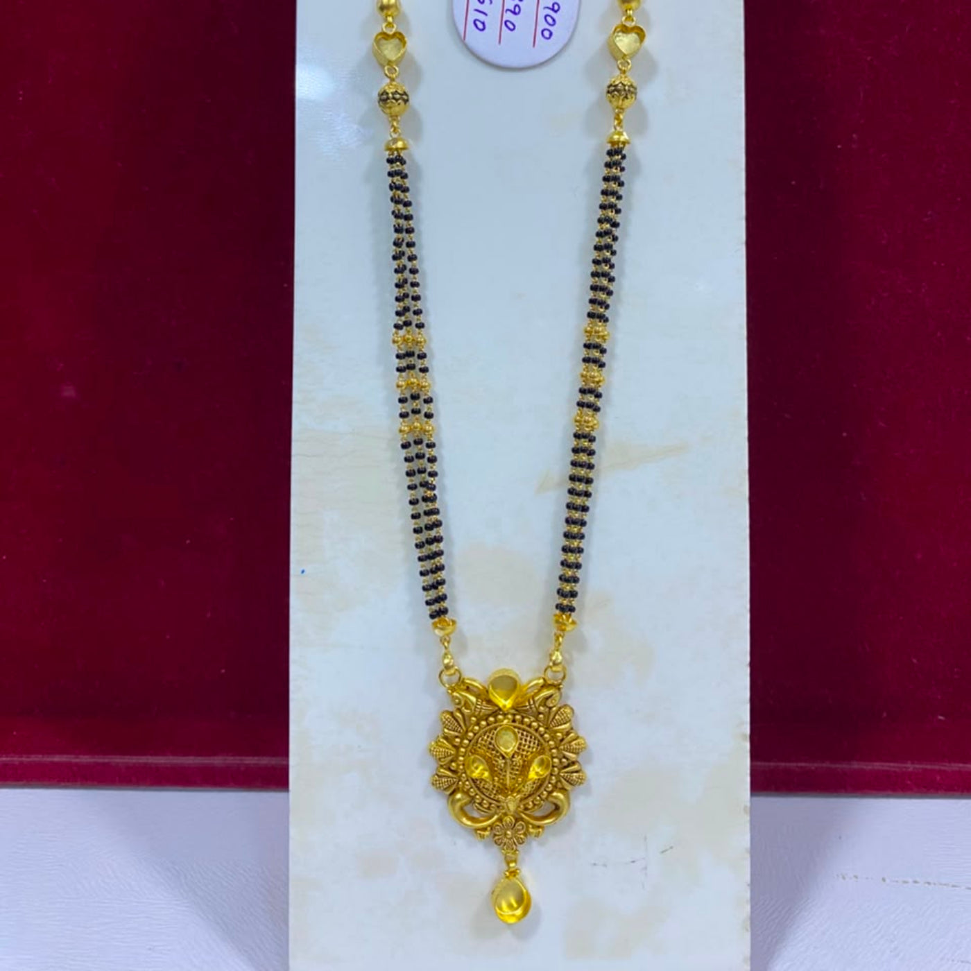 A gold and black beaded necklace with a decorative gold pendant, displayed on a white jewelry stand against a dark red background. A white price tag is attached near the top.