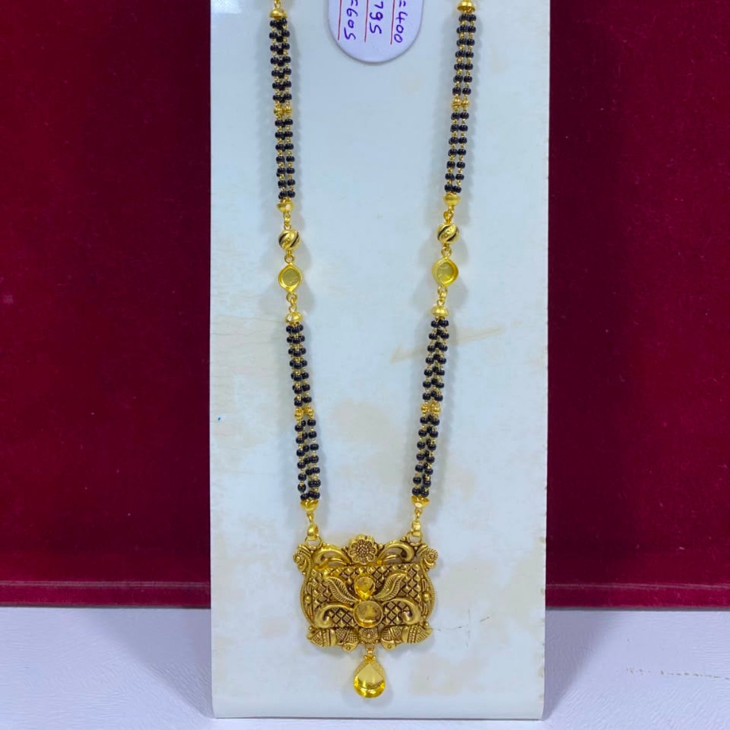 A traditional Indian gold and black beaded necklace with a detailed gold pendant, displayed on a white marble stand against a dark red background.