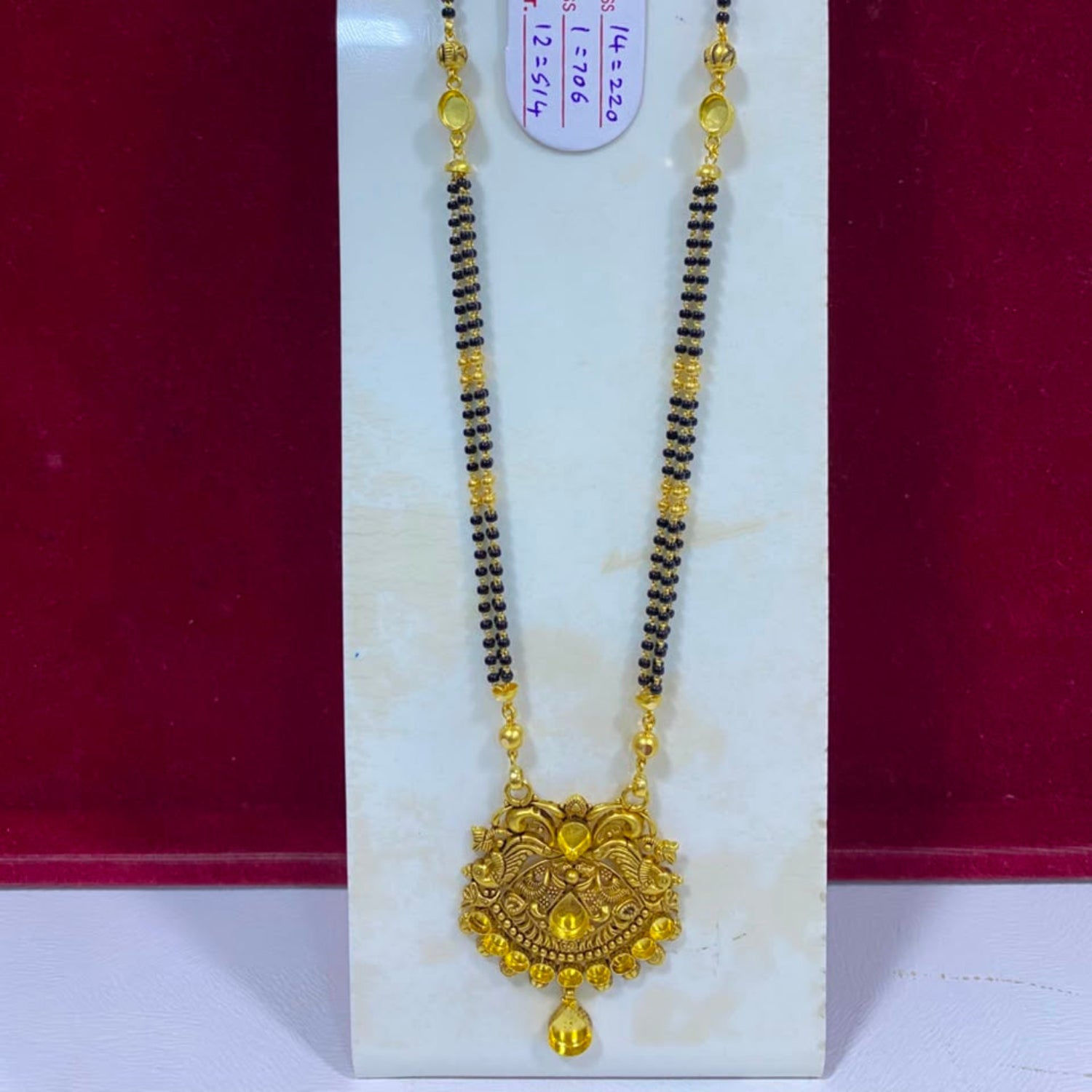 A traditional Indian gold and black beaded necklace with a detailed gold pendant, displayed on a white stand against a dark red background.