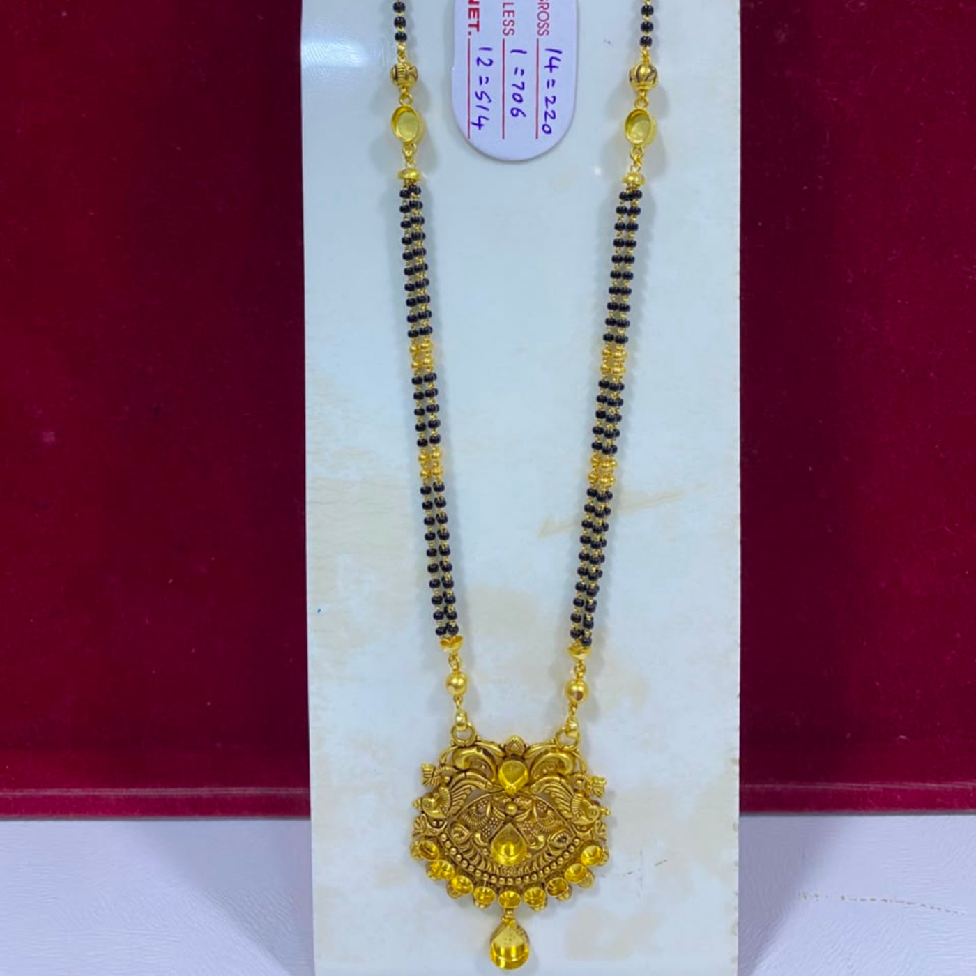A traditional Indian gold and black beaded necklace with a detailed gold pendant, displayed on a white stand against a dark red background.