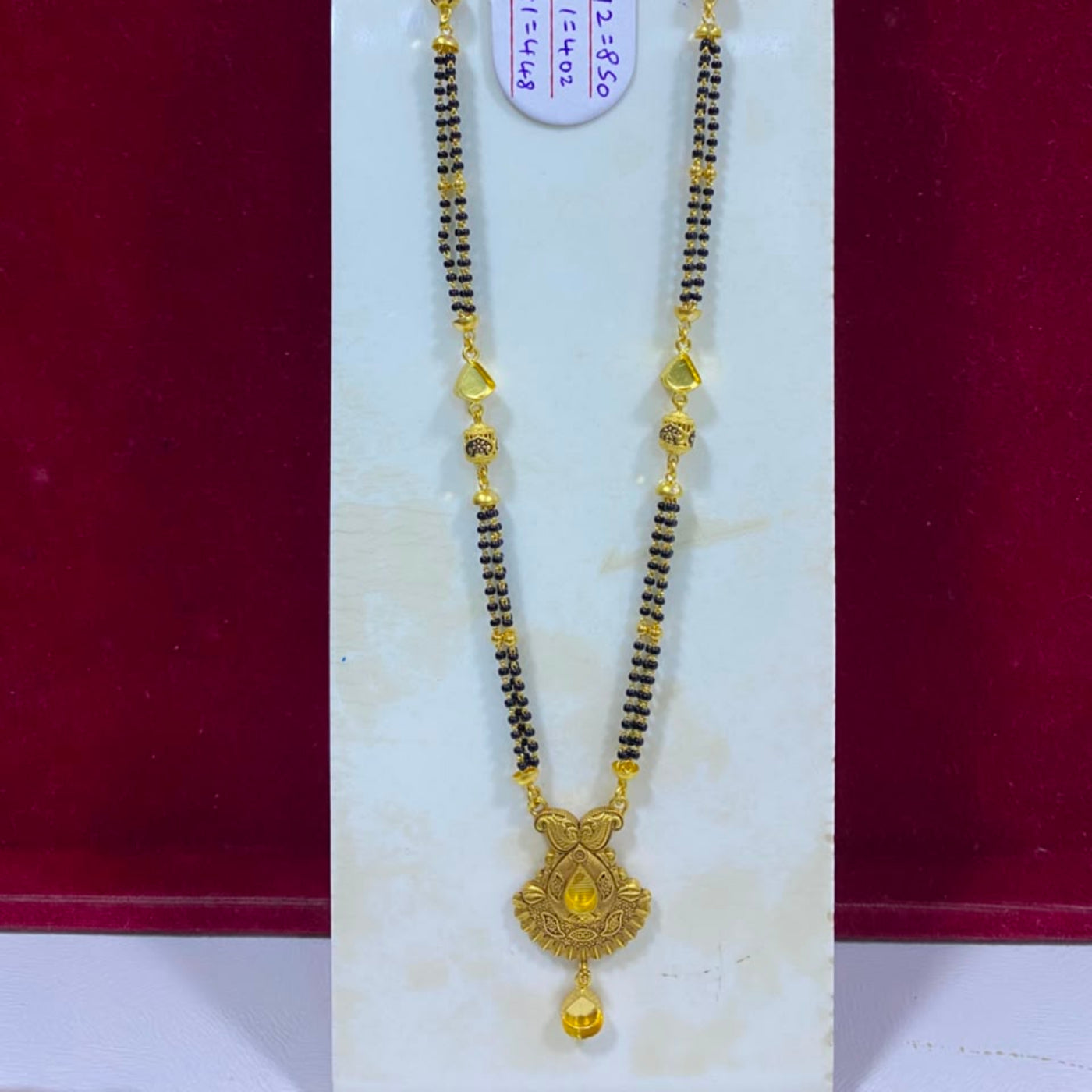 A traditional Indian gold and black beaded necklace (Murugai) with a detailed gold pendant, displayed on a white jewelry stand against a dark red background.