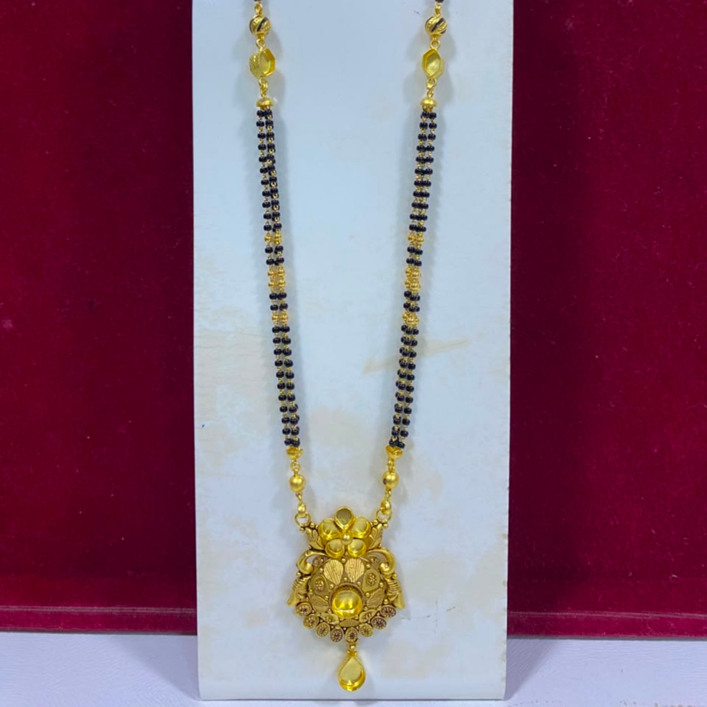A traditional Indian gold-plated necklace with a black bead chain and an ornate gold pendant, displayed on a white stand against a dark red background.