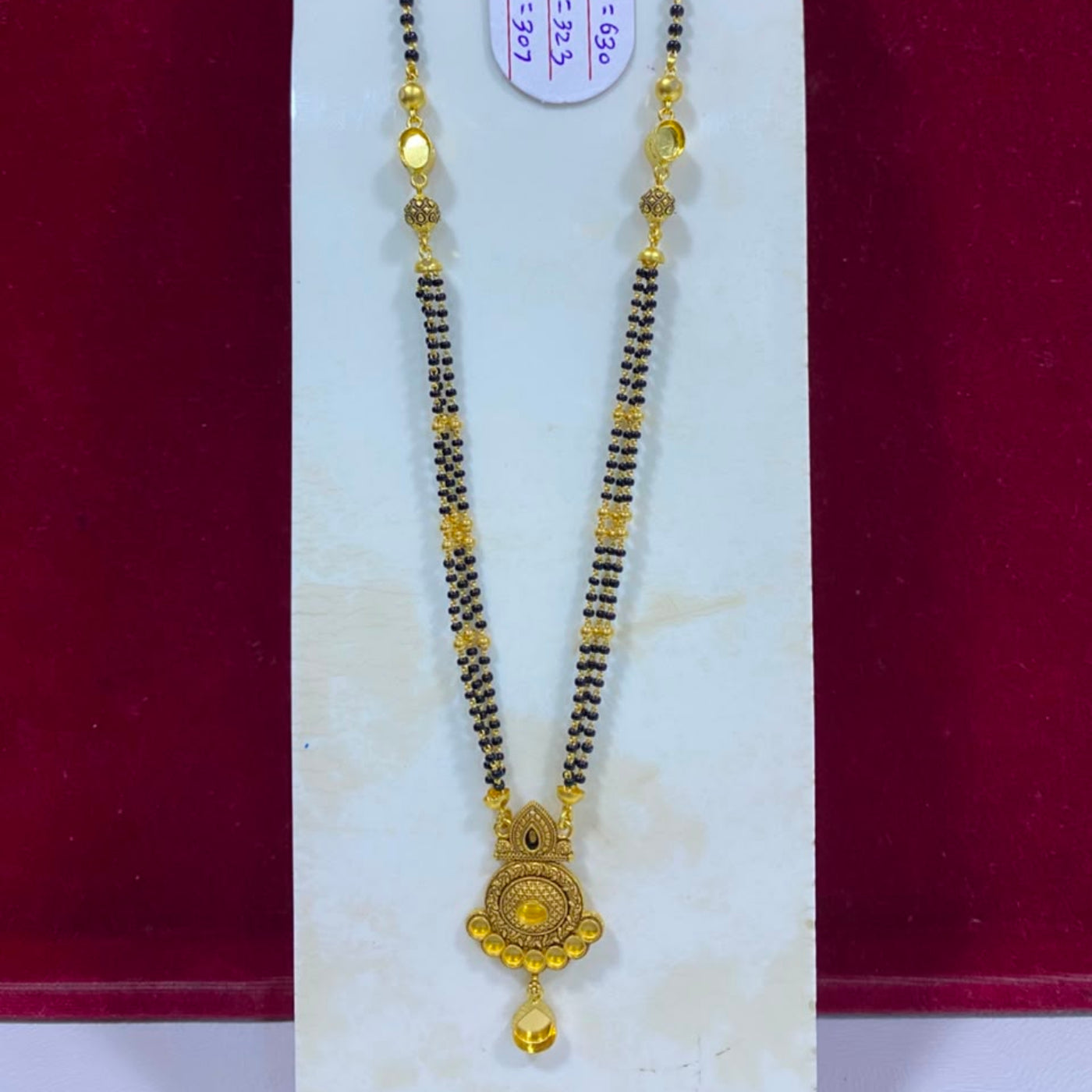 A traditional Indian gold and black beaded necklace (Magga) with a detailed gold pendant, displayed on a white jewelry stand against a dark red background.