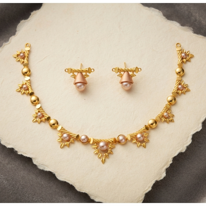 Gold-toned jewelry set with a necklace and matching earrings, featuring floral motifs, small pink gemstones, and round pearl-like beads, arranged on a textured off-white stone slab over a dark gray fabric background.