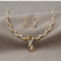 Gold-toned jewelry set featuring a necklace and matching earrings with ribbon-shaped pendants and small square accents, displayed on a soft beige fabric background.