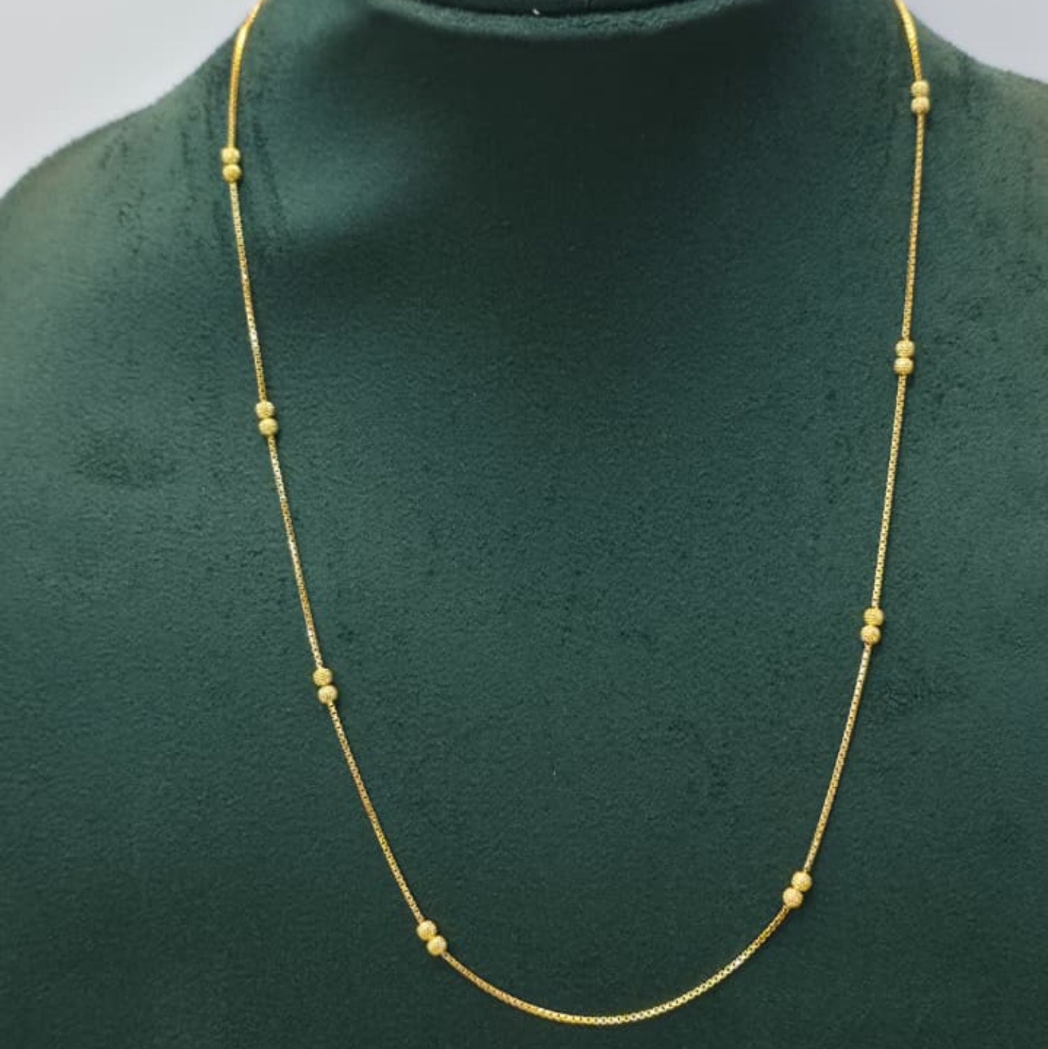 A delicate gold chain necklace with small bead-like spaced accents, displayed on a dark green velvet jewelry bust against a light gray background.