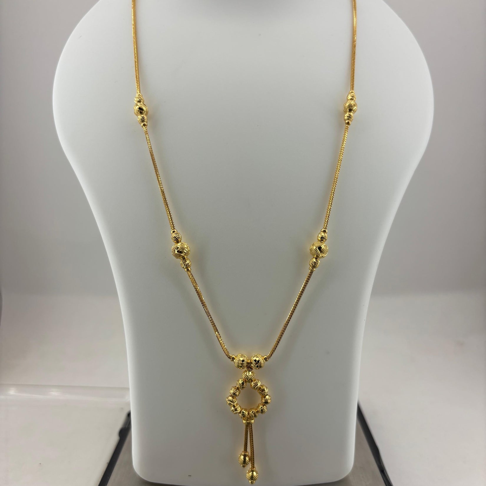 A gold necklace displayed on a white jewelry bust, with a digital scale visible underneath. The necklace features a delicate chain with small bead details and a central key-shaped pendant.