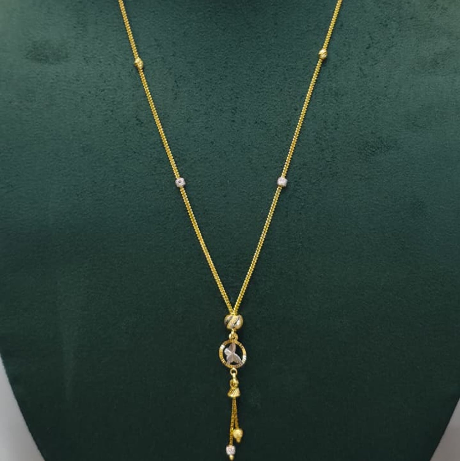 A gold chain necklace with a circular pendant and dangling tassels, displayed on a dark green velvet jewelry bust against a light gray background.