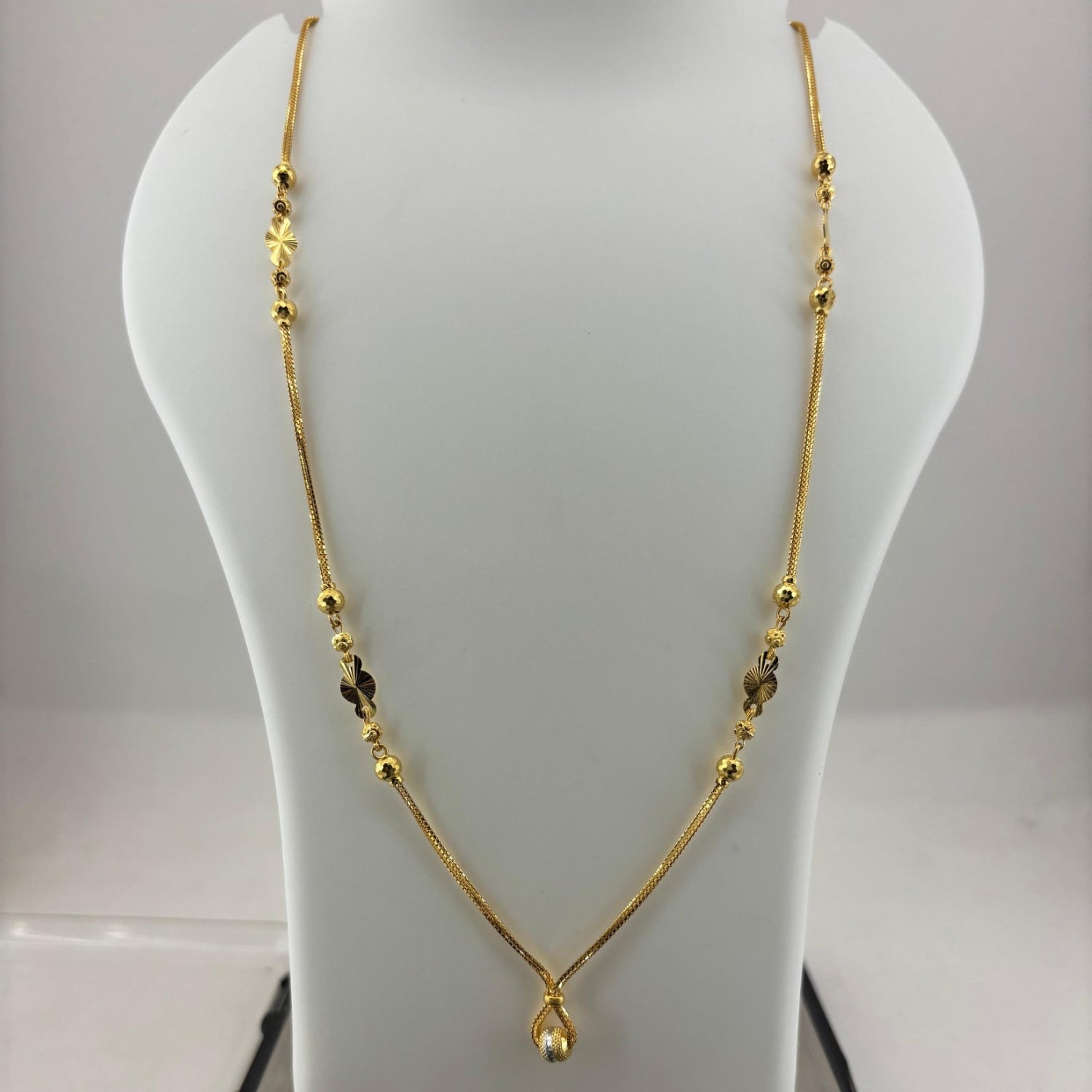 A gold necklace with small bead details and a teardrop pendant, displayed on a white jewelry bust with a digital scale underneath.