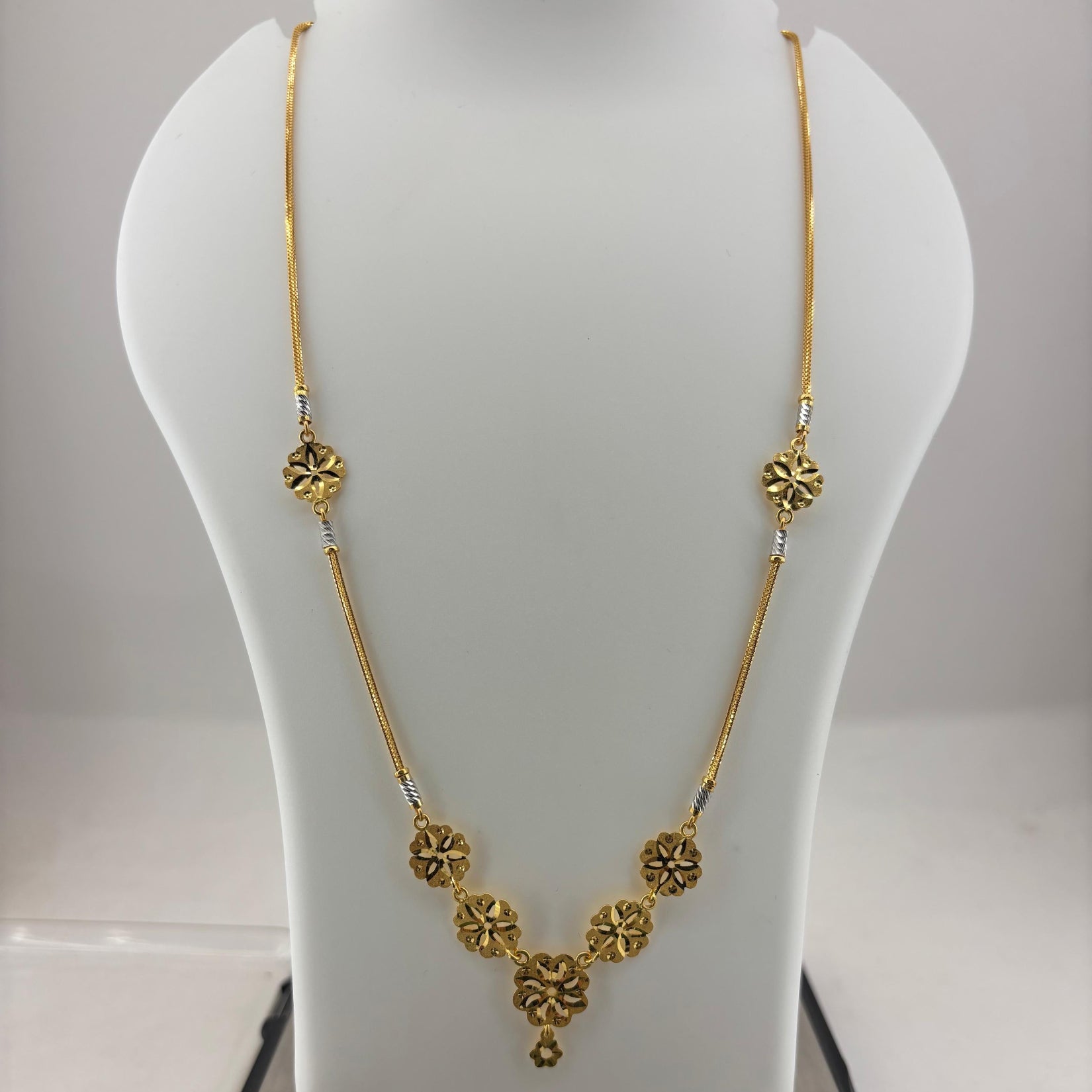 A gold necklace displayed on a white mannequin bust, with a digital scale visible underneath. The necklace features a delicate chain with ornate circular floral motifs and a central pendant.
