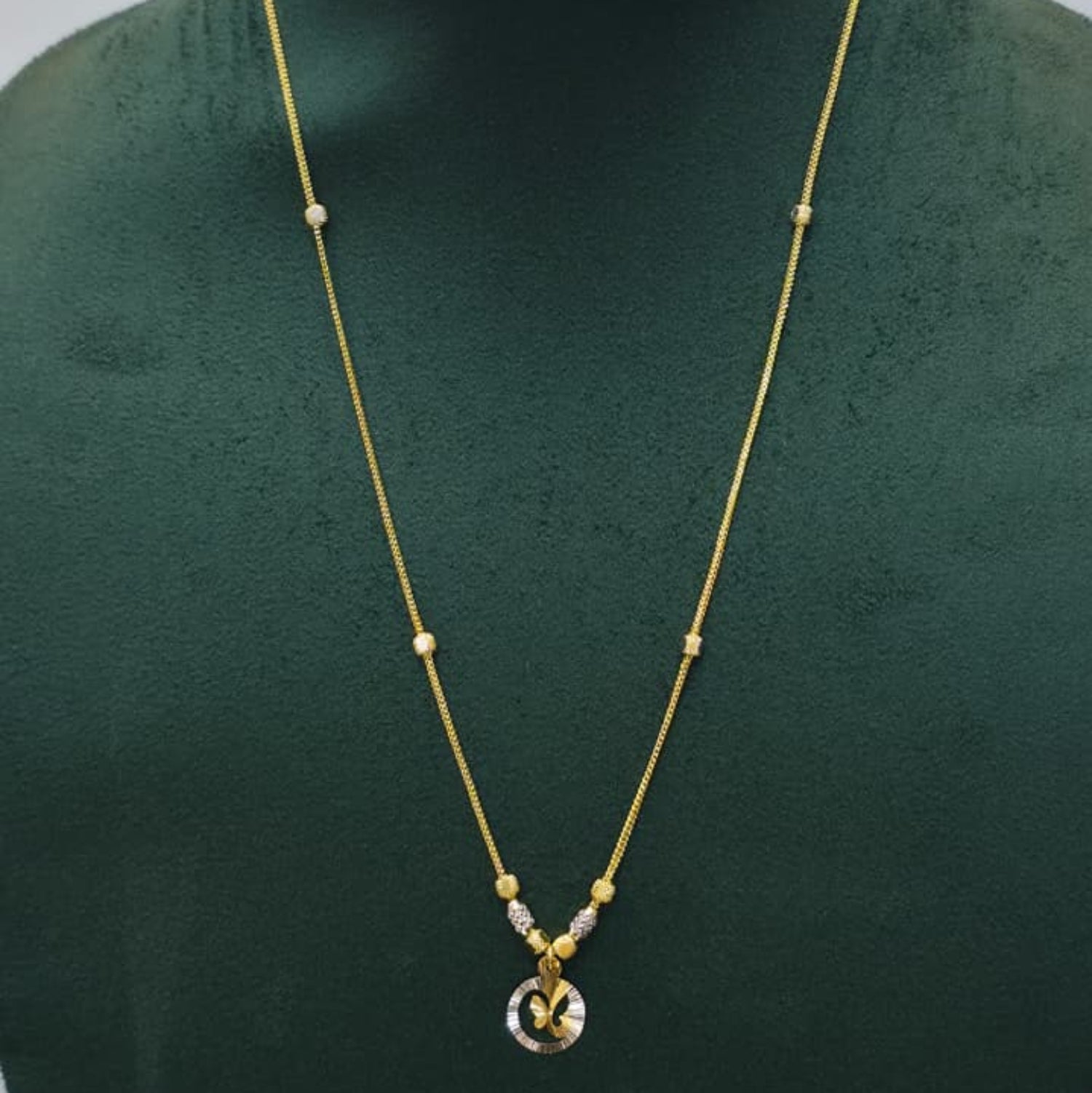 A delicate gold chain necklace with small bead accents and a round pendant, displayed on a dark green jewelry bust against a light gray background.