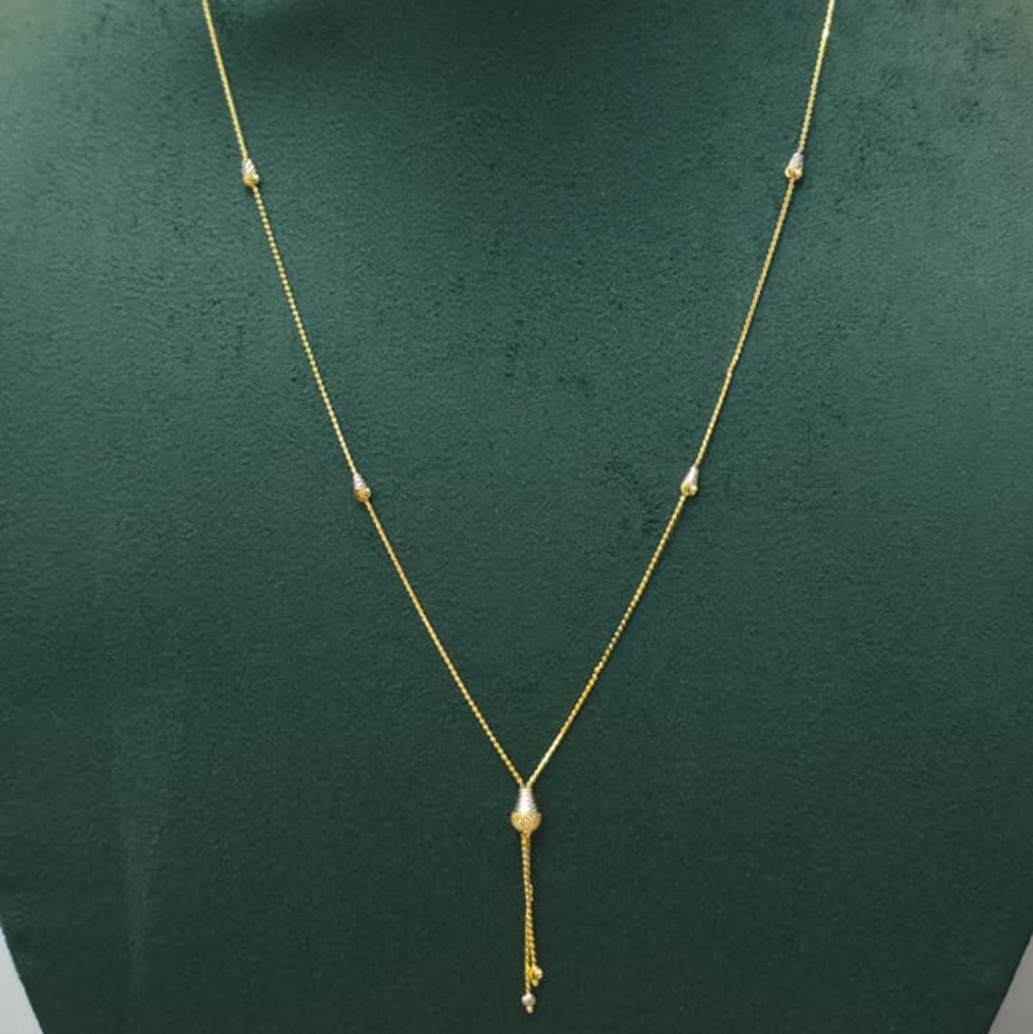 A delicate gold necklace with a central teardrop-shaped pearl and a dangling gold charm, displayed on a dark green jewelry bust.