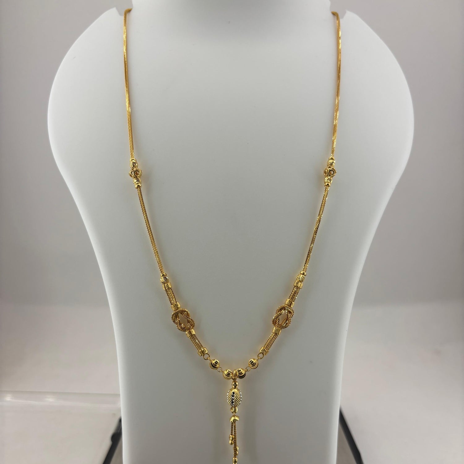 A gold necklace displayed on a white jewelry bust, with a digital scale visible underneath. The necklace features a textured chain with small circular and oval links and a dangling pendant.