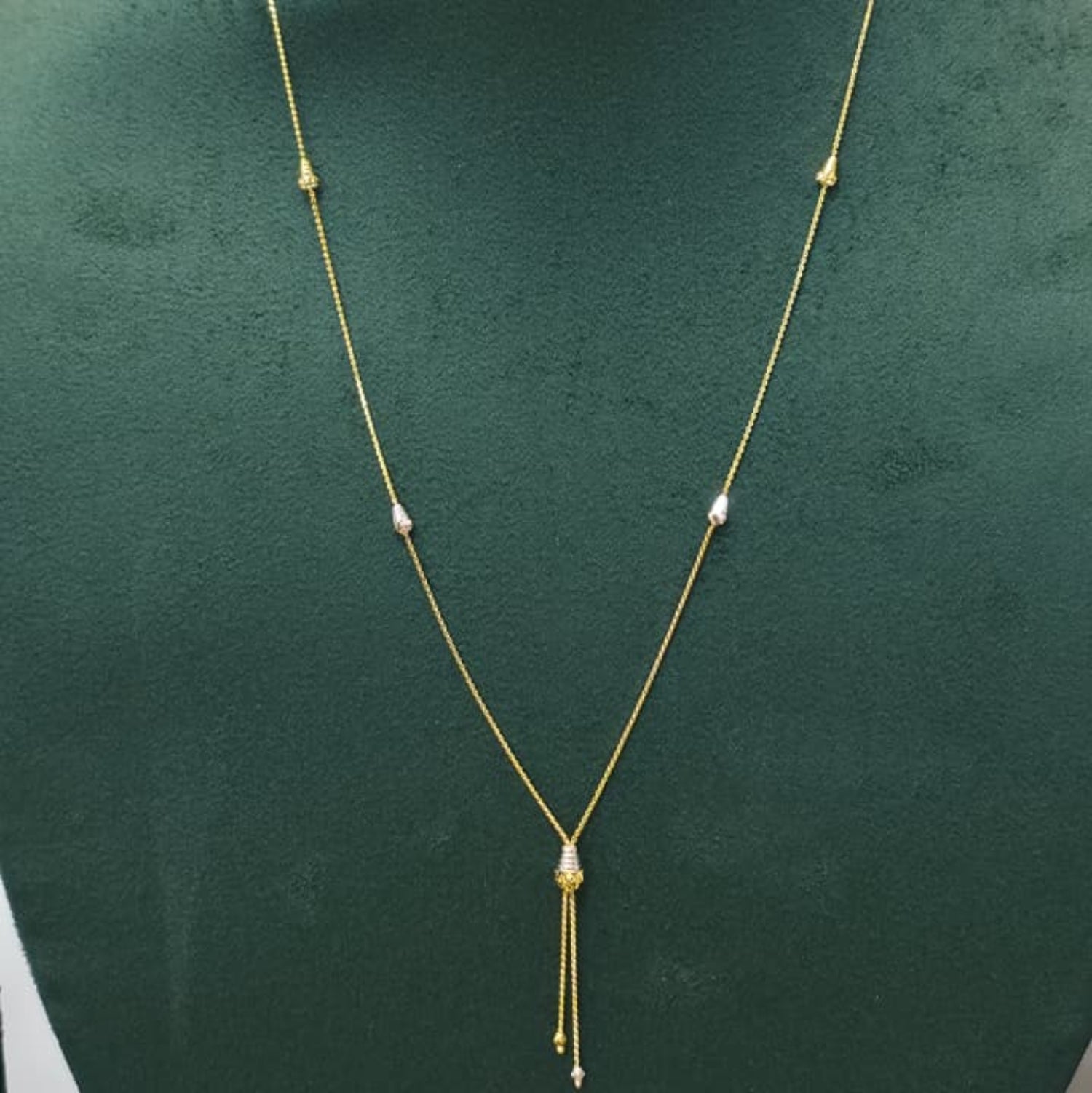 A delicate gold necklace displayed on a dark green jewelry bust. The necklace features small spaced gold beads, a central teardrop-shaped gold bead, and two short dangling chains at the bottom with tiny clear stones.
