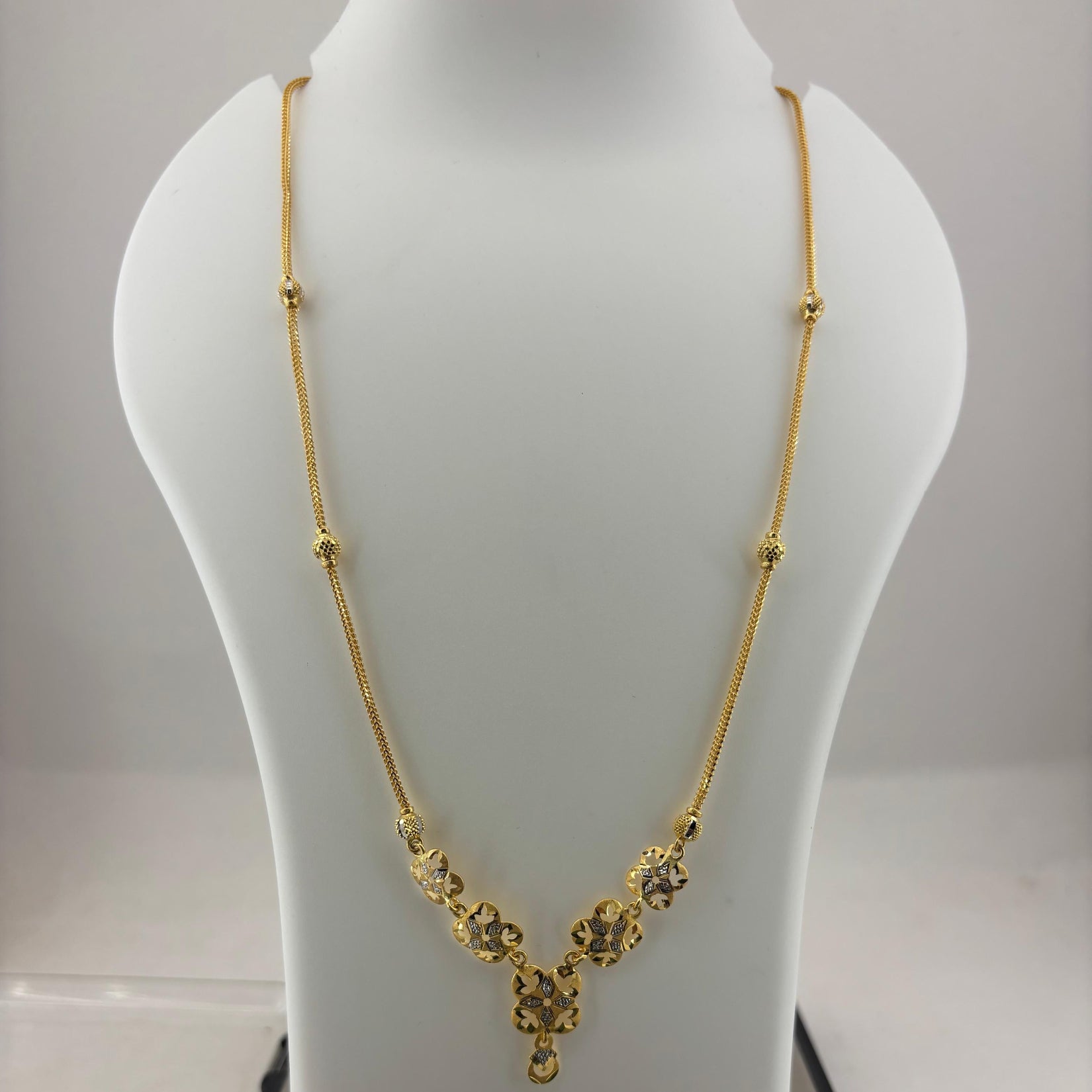 A gold necklace displayed on a white mannequin bust, with a digital scale visible underneath. The necklace features a delicate chain with small bead accents and a V-shaped pendant with floral clusters and a teardrop charm.