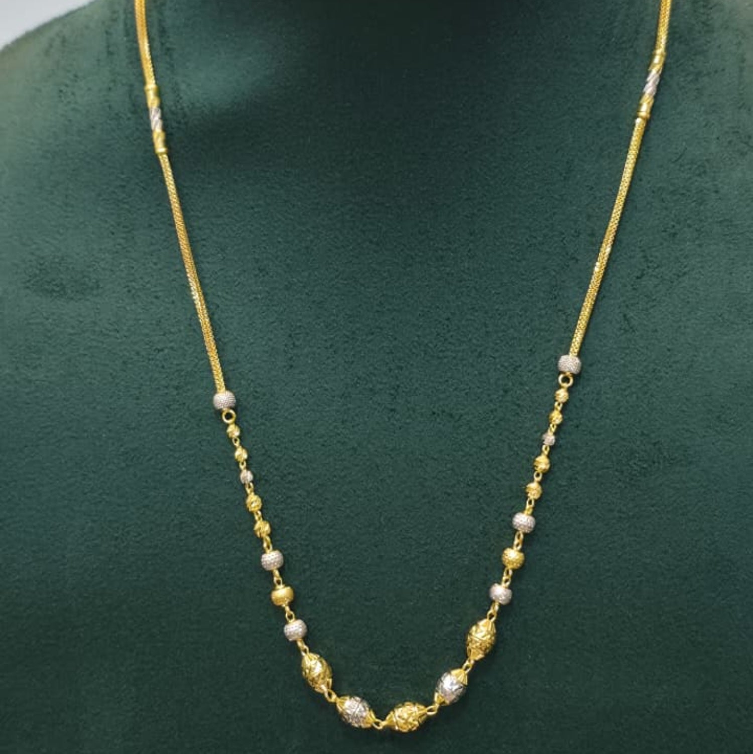 A gold chain necklace with small white bead accents displayed on a dark green jewelry bust.