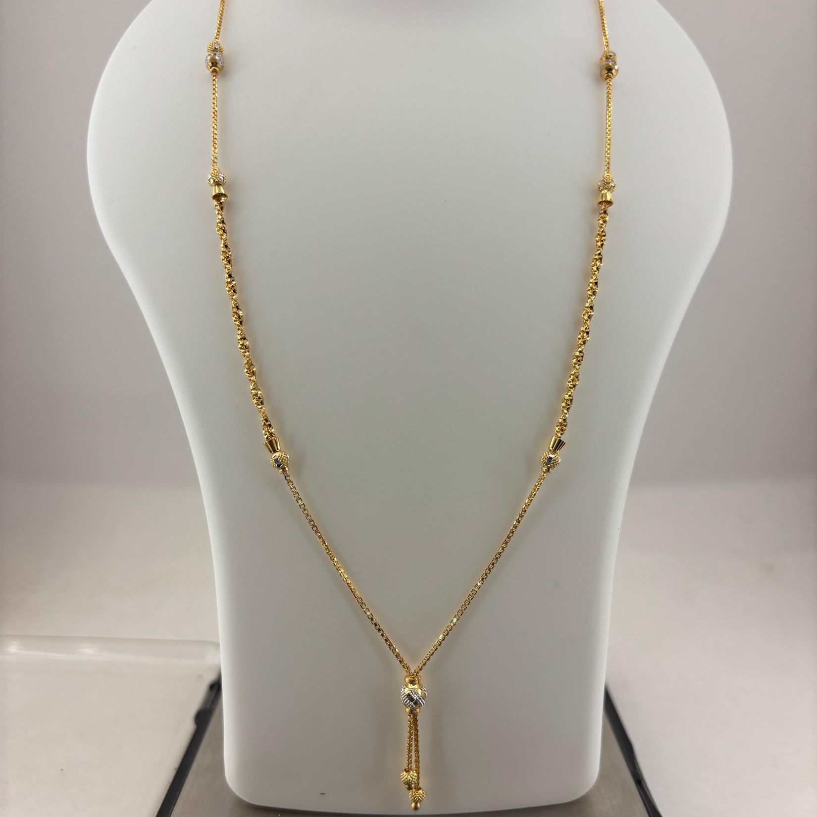 A gold necklace displayed on a white jewelry bust, with a digital scale visible underneath. The necklace has a delicate chain with small bead details and a central pendant with a clear stone.