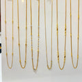 Nine gold-tone chain necklaces with various bead and spacer details are displayed on a white vertical rack with price tags attached, set against a softly blurred indoor background.